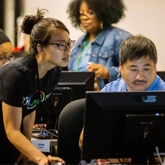 A woman with glasses and her hair in a bun leans over to assist a man sitting at a computer in a crowded workspace. She wears a black t-shirt with a "GoogleServe" logo and an "Erika" name tag. Other people are visible working at computers in the background.