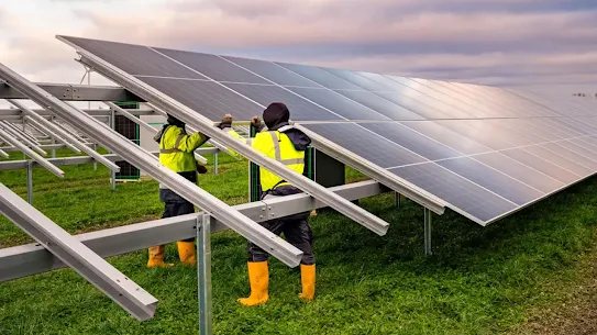 Dua orang yang memakai bot oranye dan rompi kuning memasang panel surya untuk pusat data di Fredericia, Denmark.