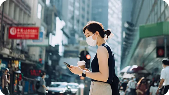 A woman wearing a face mask looks at her phone while walking on a busy city street.