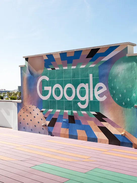A rooftop with the Google logo on a wall. Palm trees and boats are in the distance.