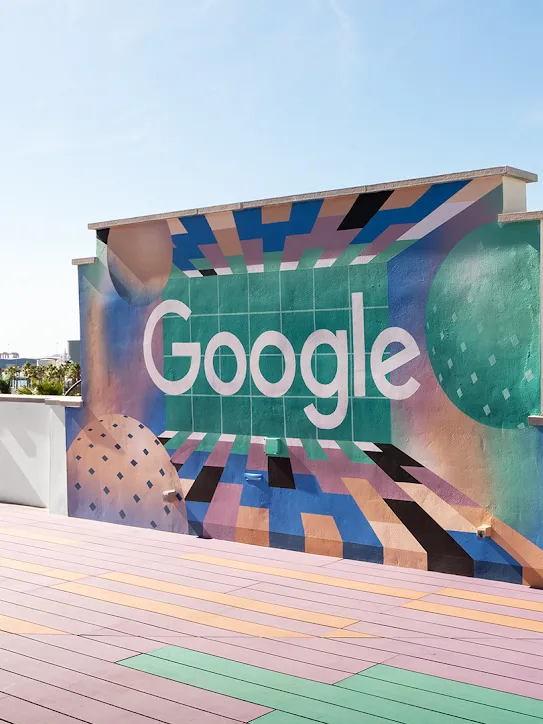 Atap gedung dengan logo Google di dinding. Pohon palem dan sejumlah kapal tampak di kejauhan.