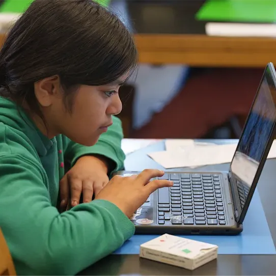 Young girl in green sweatshirt sits at desk with laptop open while she stares and starts to type