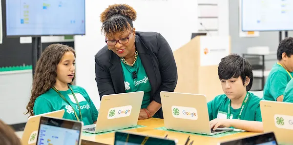 An adult helps teenagers use a visual programming tool on laptops in a classroom