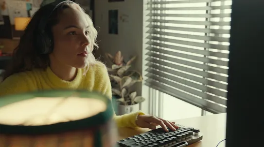 Sarı kazak giymiş kulaklık takan bir kişi klavyede bir şeyler yazıyor. Ön tarafta bir lamba görünüyor.