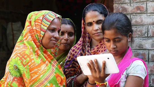 A young person uses a device, while older people stand behind her, watching closely with caring expressions.