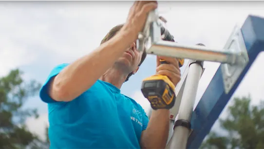 Man with a blue shirt on using a yellow power drill to work on a piece of steel that is largely out of the frame