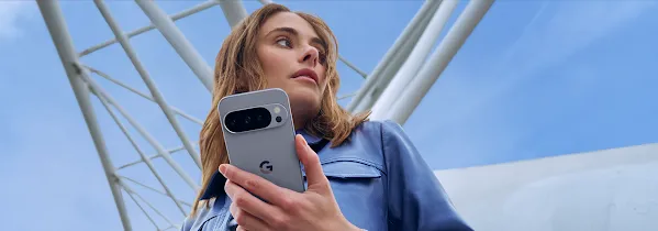 Mavi ceketli bir kişi, alçak bir açıdan çekilen fotoğrafta gri Pixel telefon tutuyor. Arka planda parlak mavi bir gökyüzü ve beyaz mimari kirişler var.