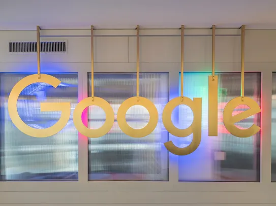 Le logo Google est suspendu à des tiges métalliques devant un mur semi-transparent, éclairé en bleu, rose et turquoise.
