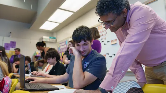 Male teacher reading a page with student looking at same paper at desk in a class room full of students and faculty 
