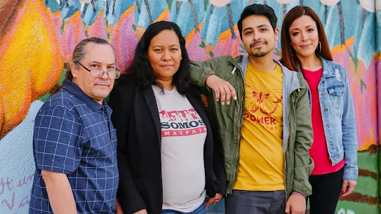 Four individuals in variously colored shirts and t-shirts stand in front of a colorful mural that appears to depict people and abstract shapes.