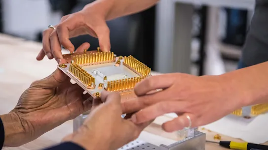 Seseorang menggunakan dua tangan untuk menyerahkan prosesor kuantum kepada orang lain, yang menerimanya dengan dua tangan.