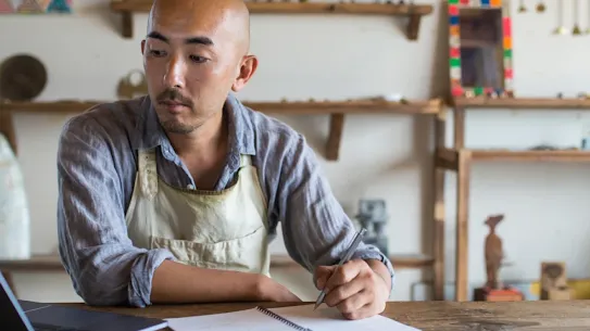 Seseorang yang mengenakan celemek kerja duduk di depan meja. Laptopnya terbuka dan dia memegang pena untuk mencatat.