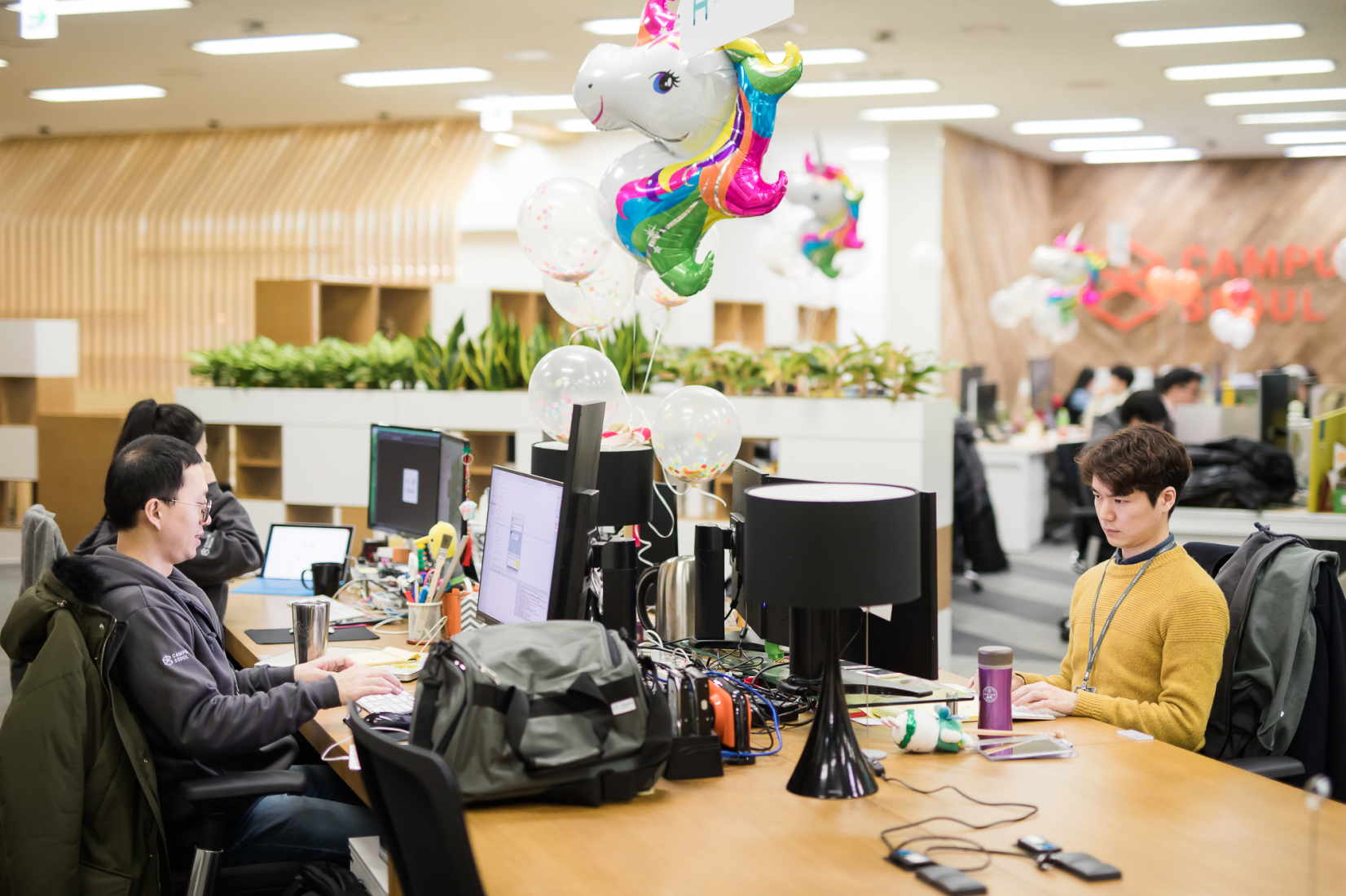 한 무리의 젊은 사람들이 현대적인 사무실에서 일하고 있고 책상 위에 무지개 유니콘 풍선 다발이 놓인 모습
