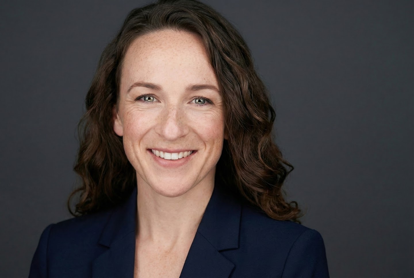 A woman with freckles and brown hair smiles warmly against a dark gray background. She is wearing a professional navy blue blazer in a clean, high-resolution portrait.