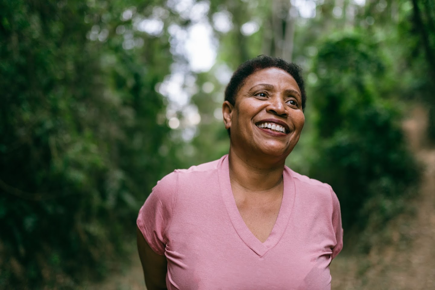 Woman in pink shirt smiles while standing in the forest.